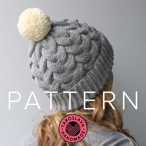 Op de afbeelding: Grijze gebreide beanie met een witte pompon bovenop. De beanie heeft een kabelbreipatroon en de tekst "PATTERN" staat in witte letters onderaan de afbeelding.