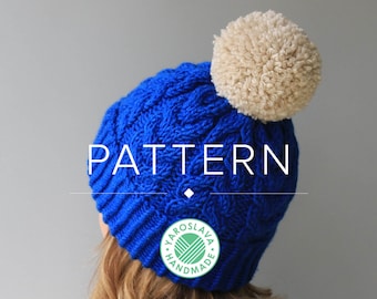 May include: Blue knitted hat with a white pom pom and a cable knit pattern. The hat has a label that says "Yaroslava Handmade".