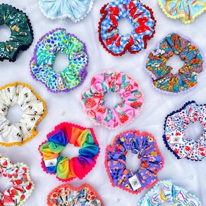 May include: A collection of thirteen fabric hair scrunchies featuring various patterns including rainbows, strawberries, cherries, bees, and floral prints. Each scrunchie is finished with a decorative pom-pom trim along the outer edge.