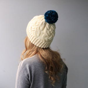 May include: A white knitted beanie with a blue pom pom on top. The beanie has a cable knit pattern.