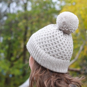 May include: A gray knitted beanie with a large pom pom on top. The beanie has a waffle stitch pattern.