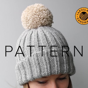 May include: A gray knitted beanie with a white pom pom on top. The beanie has a ribbed texture and the word "PATTERN" is written in black letters across the front. The beanie is worn by a person with blonde hair and the text "YAROSLAVA HANDMADE" is visible in the upper right corner of the image.