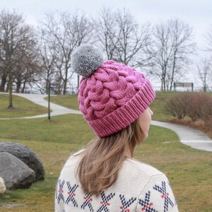 K&ouml;nnte beinhalten: Eine rosa Strickm&uuml;tze mit einem grauen Bommel. Die M&uuml;tze hat ein Zopfmuster und wird von einer Person mit langen braunen Haaren getragen. Die Person tr&auml;gt einen wei&szlig;en Pullover mit einem rot-blauen Muster.
