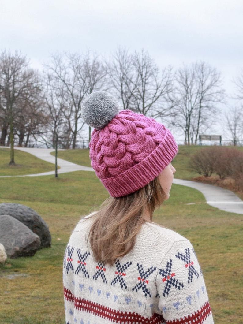 K&ouml;nnte beinhalten: Eine rosa Strickm&uuml;tze mit einem grauen Bommel. Die M&uuml;tze hat ein Zopfmuster und wird von einer Person mit langen braunen Haaren getragen. Die Person tr&auml;gt einen wei&szlig;en Pullover mit einem rot-blauen Muster.