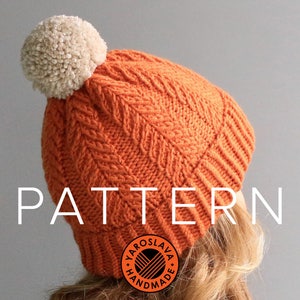 Op de afbeelding: Oranje gebreide muts met een witte pompon en een kabelbreipatroon. De muts heeft een label met de tekst "Yaroslava Hand Made".