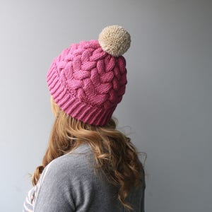 K&ouml;nnte beinhalten: Eine rosa gestrickte M&uuml;tze mit einem wei&szlig;en Bommel oben. Die M&uuml;tze hat ein Zopfmuster.