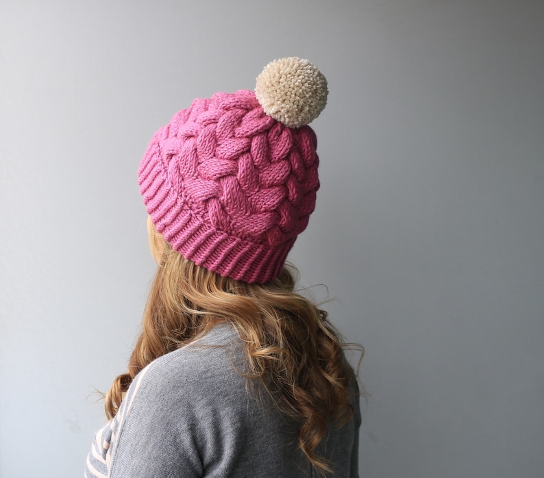 K&ouml;nnte beinhalten: Eine rosa gestrickte M&uuml;tze mit einem wei&szlig;en Bommel oben. Die M&uuml;tze hat ein Zopfmuster.