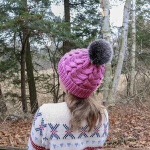 K&ouml;nnte beinhalten: Eine rosa gestrickte M&uuml;tze mit einem geflochtenen Muster und einem flauschigen grauen Bommel. Die M&uuml;tze wird von einer Person getragen, die in einem Waldgebiet steht.
