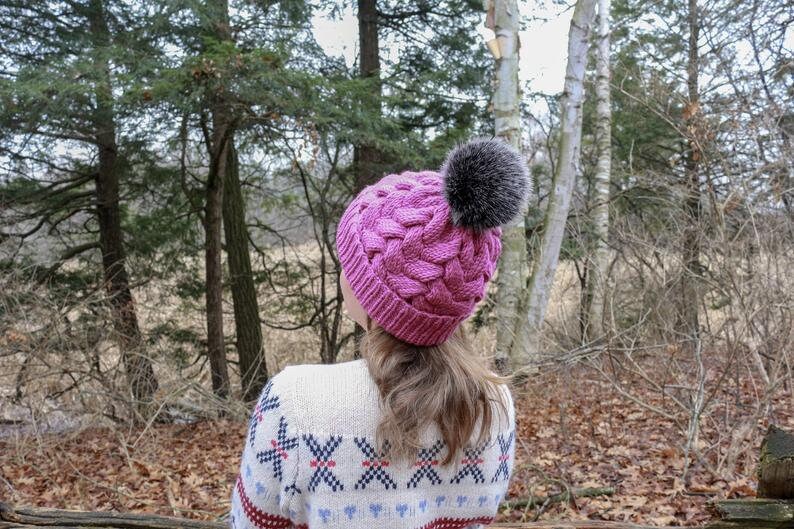 K&ouml;nnte beinhalten: Eine rosa gestrickte M&uuml;tze mit einem geflochtenen Muster und einem flauschigen grauen Bommel. Die M&uuml;tze wird von einer Person getragen, die in einem Waldgebiet steht.