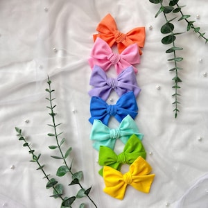 Könnte beinhalten: Ein vertikaler Stapel aus sieben Haarspangen aus Stoff in einer lebendigen Regenbogenfarbpalette, darunter Orange, Pink, Lavendel, Blau, Türkis, Limettengrün und Gelb. Jede Schleife verfügt über ein geknotetes Mittelstück und eine matte Oberfläche.