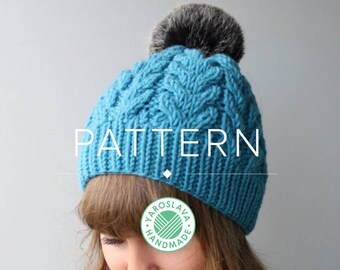 May include: A blue knitted hat with a cable knit pattern and a faux fur pom pom. The hat is worn by a person with brown hair and is labeled "PATTERN".