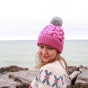 K&ouml;nnte beinhalten: Eine rosa Strickm&uuml;tze mit Zopfmuster und einem grauen Bommel. Die M&uuml;tze wird von einer Person getragen, die vor einem Gew&auml;sser steht.