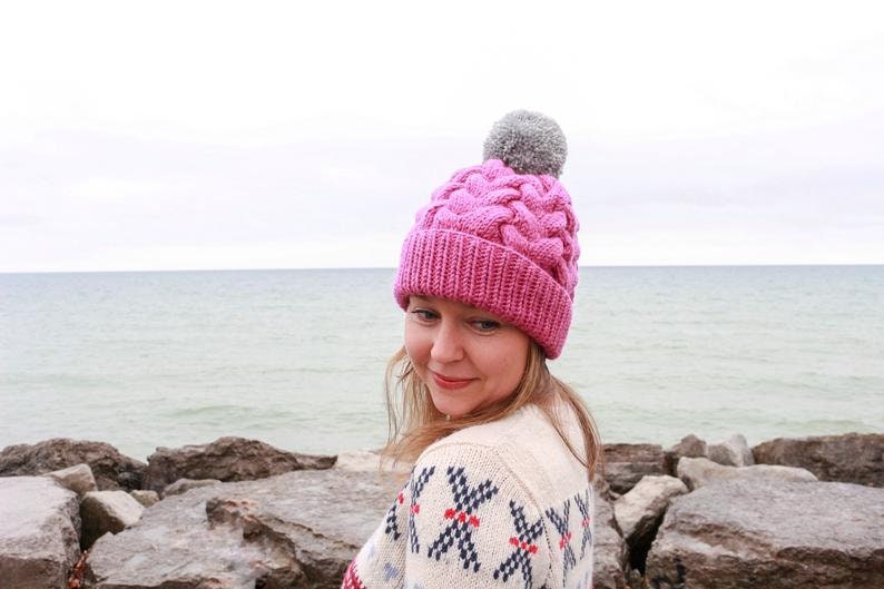 K&ouml;nnte beinhalten: Eine rosa Strickm&uuml;tze mit Zopfmuster und einem grauen Bommel. Die M&uuml;tze wird von einer Person getragen, die vor einem Gew&auml;sser steht.
