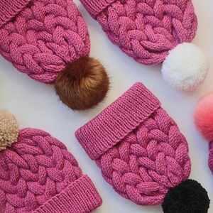 K&ouml;nnte beinhalten: Eine Sammlung von rosa gestrickten M&uuml;tzen mit verschiedenen farbigen Bommeln. Die M&uuml;tzen haben ein Zopfmuster und sind aus weichem, flauschigem Garn gefertigt.