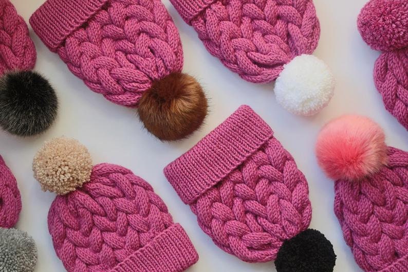 K&ouml;nnte beinhalten: Eine Sammlung von rosa gestrickten M&uuml;tzen mit verschiedenen farbigen Bommeln. Die M&uuml;tzen haben ein Zopfmuster und sind aus weichem, flauschigem Garn gefertigt.
