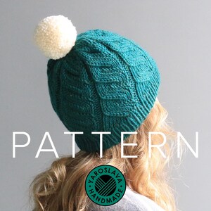 May include: A teal blue knitted hat with a white pom pom on top. The hat has a cable knit pattern and the text "PATTERN" is written in white letters across the bottom of the image.