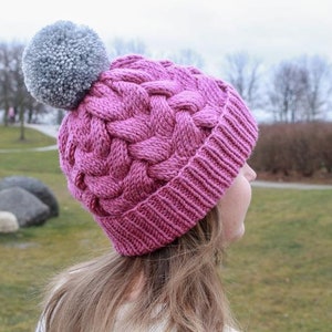 K&ouml;nnte beinhalten: Eine rosa Strickm&uuml;tze mit einem geflochtenen Muster und einem grauen Bommel oben. Die M&uuml;tze wird von einer Person mit langen braunen Haaren getragen.