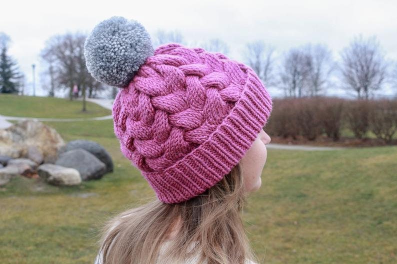 K&ouml;nnte beinhalten: Eine rosa Strickm&uuml;tze mit einem geflochtenen Muster und einem grauen Bommel oben. Die M&uuml;tze wird von einer Person mit langen braunen Haaren getragen.