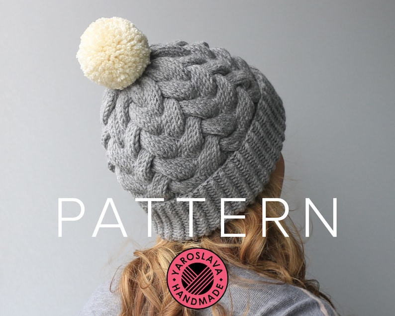 K&ouml;nnte beinhalten: Graue Strickm&uuml;tze mit einem wei&szlig;en Bommel oben. Die M&uuml;tze hat ein Zopfmuster und der Text "PATTERN" ist in wei&szlig;en Buchstaben am unteren Rand des Bildes geschrieben.