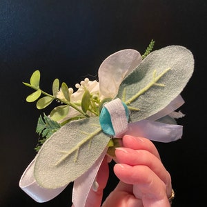 Pode incluir: Um boutonniere floral branco e verde com uma fita branca e um bot&atilde;o turquesa.