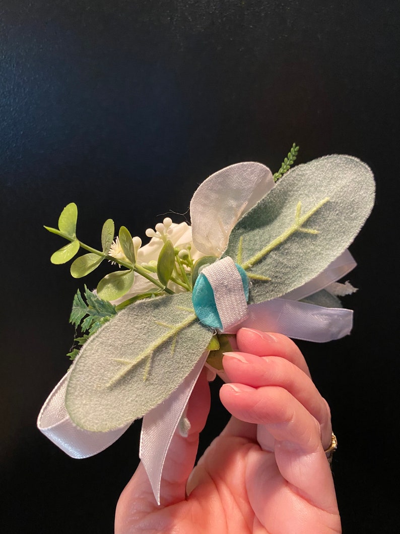 Pode incluir: Um boutonniere floral branco e verde com uma fita branca e um bot&atilde;o turquesa.