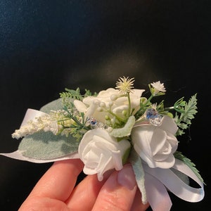 Pode incluir: Uma boutonniere de flores brancas com uma fita de cetim branca e detalhes de strass azuis.