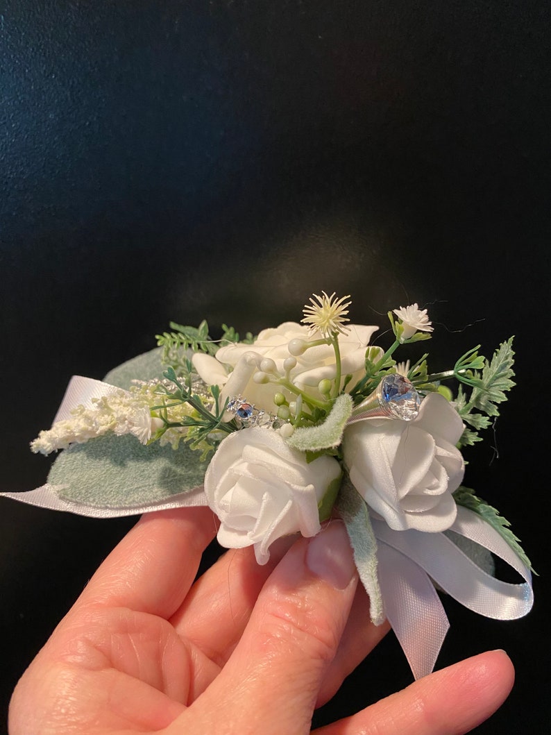 Pode incluir: Uma boutonniere de flores brancas com uma fita de cetim branca e detalhes de strass azuis.