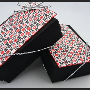 May include: Two black gift boxes with lids, both wrapped in white paper with a repeating pattern of red circles, black circles, and the word "MOM" in black. The boxes are tied with silver ribbon.