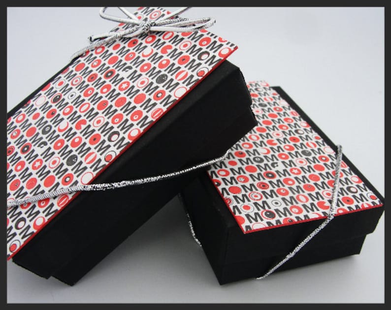 May include: Two black gift boxes with lids, both wrapped in white paper with a repeating pattern of red circles, black circles, and the word "MOM" in black. The boxes are tied with silver ribbon.