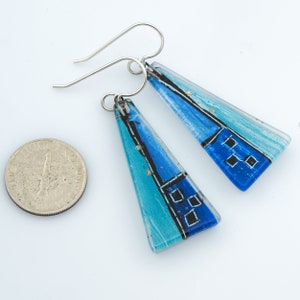 May include: A pair of blue and turquoise glass earrings with silver hooks. The glass is painted with black lines and white squares. The earrings are shaped like triangles.