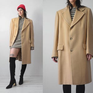 Op de afbeelding: Een beige overjas met een klassiek ontwerp. De jas heeft een reverskraag, twee voorzakken en een knoopsluiting. Het model draagt een rode muts, een zwart-wit gestreepte jurk en zwarte over-the-knee laarzen.