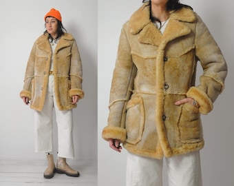 May include: A tan shearling coat with a button-up front, large pockets, and a wide collar. The coat has a light brown interior and trim. The coat is worn with white trousers and a bright orange beanie.