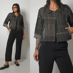 May include: A charcoal gray cropped jacket featuring three-quarter length sleeves with ribbed cuffs, a rounded collar, and a button-front closure. The fabric displays a textured grid pattern with a vertical red stitched line and two side pockets.