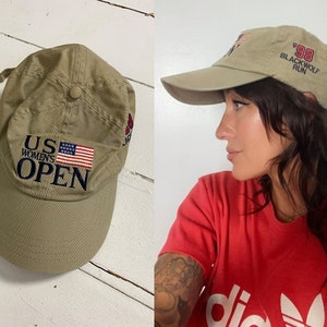 Op de afbeelding: Twee kaki baseballpetten. Eén heeft de tekst "US Women's Open" met een kleine Amerikaanse vlag. De andere heeft de tekst "'98 Blackwolf Run".