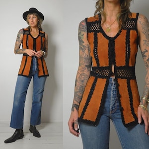 May include: A vintage suede vest in a warm orange colour, with black crochet detailing along the edges and seams. The vest has a V-neck and a button-up front. The model is wearing blue jeans and a black hat.