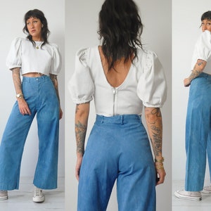 May include: A model wears a white crop top with puffed sleeves and high-waisted, wide-leg blue jeans. The top has a zipper in the back.