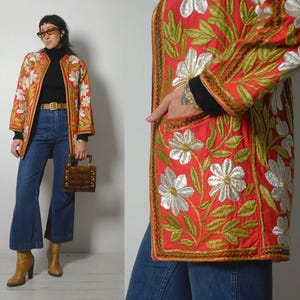 Könnte beinhalten: Rote, bestickte Jacke mit weißen und goldenen Blumenmustern. Die Jacke hat lange Ärmel und einen goldenen Besatz. Sie wird über einem schwarzen Oberteil und einer blauen Jeans getragen. Eine braune Handtasche und Stiefel vervollständigen den Look.