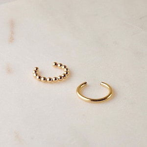 May include: Two gold ear cuffs. One ear cuff is a simple, smooth, curved shape. The other ear cuff is made of a series of small, round beads.