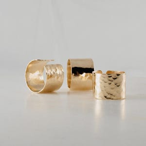 May include: Three gold-colored rings with a hammered texture are displayed on a white surface. The rings vary in size, with one open-ended and two closed, showcasing a bright, reflective finish. The jewelry is simple and elegant.
