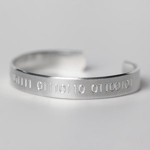 May include: A silver cuff bracelet with engraved binary code. The open-ended bracelet has a brushed metal finish and features a series of numbers etched into the surface. The bracelet is a minimalist design, suitable for everyday wear.