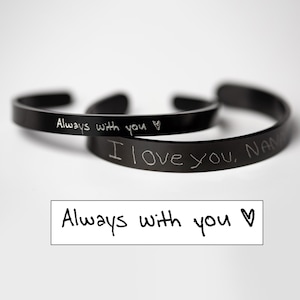 May include: Two black metal bracelets with handwritten messages. The top bracelet says "Always with you" with a heart symbol. The bottom bracelet says "I love you, NAME".