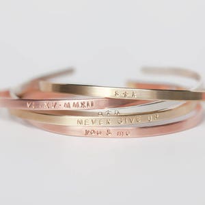 May include: Four stacked gold, silver, and rose gold bracelets with engraved messages: "S+K", "VI.KY.MMXXI", "NEVER GIVE UP", and "you & me".