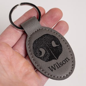 May include: An oval-shaped grey leather keychain featuring a black engraved dog nose print and the name Wilson printed in a serif font. The keychain is attached to a circular silver metal split ring and has visible stitching along the edge.