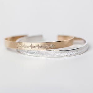 May include: Two bracelets stacked on top of each other. The top bracelet is gold and has a sound wave design engraved on it. The bottom bracelet is silver and has a textured finish.