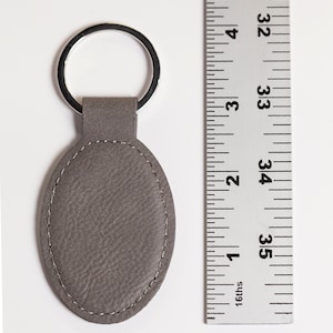 May include: An oval-shaped grey keychain with a textured surface and visible stitching along the perimeter, attached to a silver metal ring. A metal ruler is positioned next to the keychain, showing a length of approximately 2.5 inches.