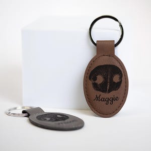 May include: Two oval-shaped brown leather keychains with metal rings. Each keychain features a black engraved dog nose print and a name printed below it. One keychain is upright, showing the name Maggie, while the other lies flat in the foreground.