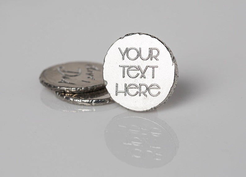 May include: Close-up of three silver-colored, coin-shaped objects. The front-most coin has the words "YOUR TEXT HERE" engraved on it. The coins have a textured, uneven edge. The objects are on a reflective surface.