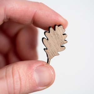 May include: A small, flat, light brown wooden cutout shaped like an oak leaf. The piece features a visible natural wood grain pattern and dark, defined edges that outline the lobed shape of the leaf, ending in a short, narrow stem at the base.