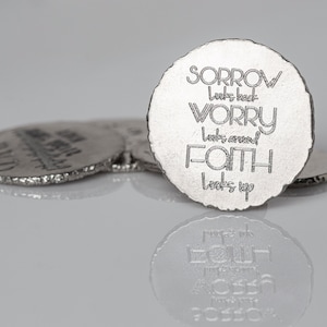May include: Silver-colored, circular coasters with rough edges. The front coaster features the words "SORROW looks back, WORRY looks around, FAITH looks up" in a stylized font. Other coasters are visible in the background, with a reflective surface.