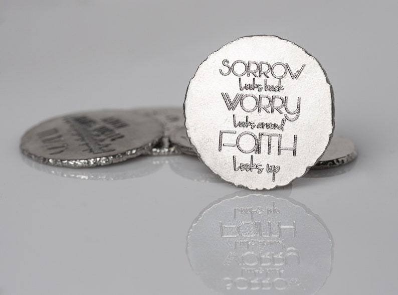 May include: Silver-colored, circular coasters with rough edges. The front coaster features the words "SORROW looks back, WORRY looks around, FAITH looks up" in a stylized font. Other coasters are visible in the background, with a reflective surface.